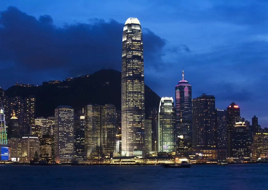 Horizonte nocturno de Hong Kong visto desde el agua, con torres de oficinas iluminadas y la silueta oscura de la montaña detrás
