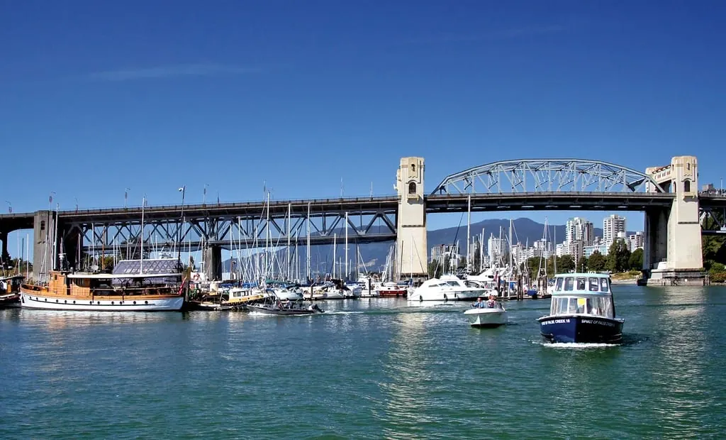 Canal em Vancouver, no Canadá, com veleiros atracados na marina e uma ponte de estrutura metálica cruzando a água, prédios altos e montanhas ao fundo