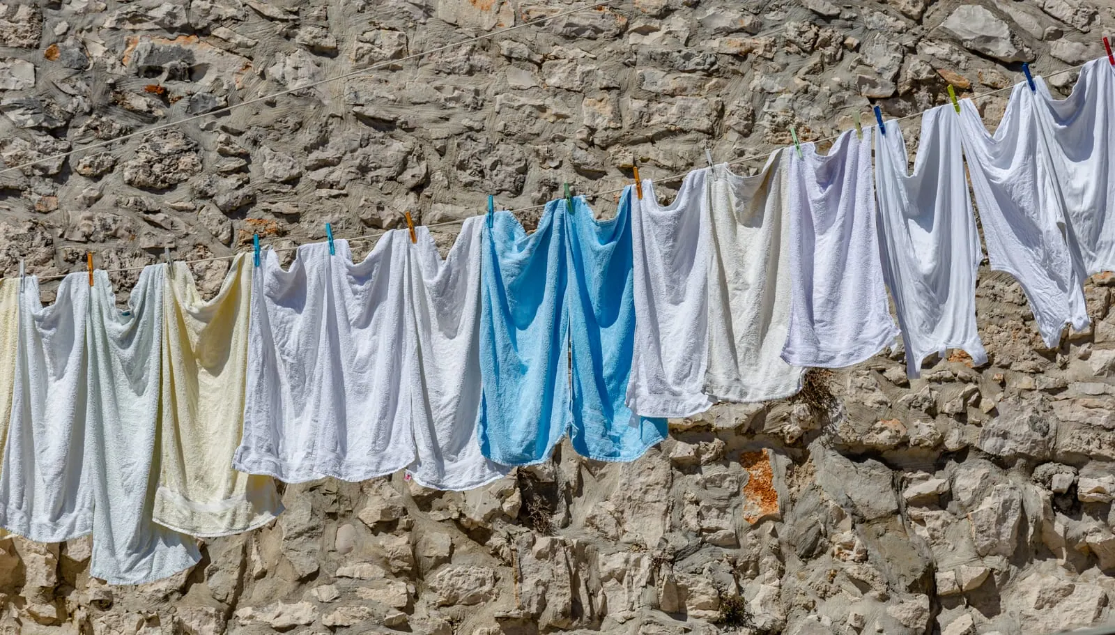 White and pale blue towels and clothes pegged along a washing line in the sun, in front of a stone wall