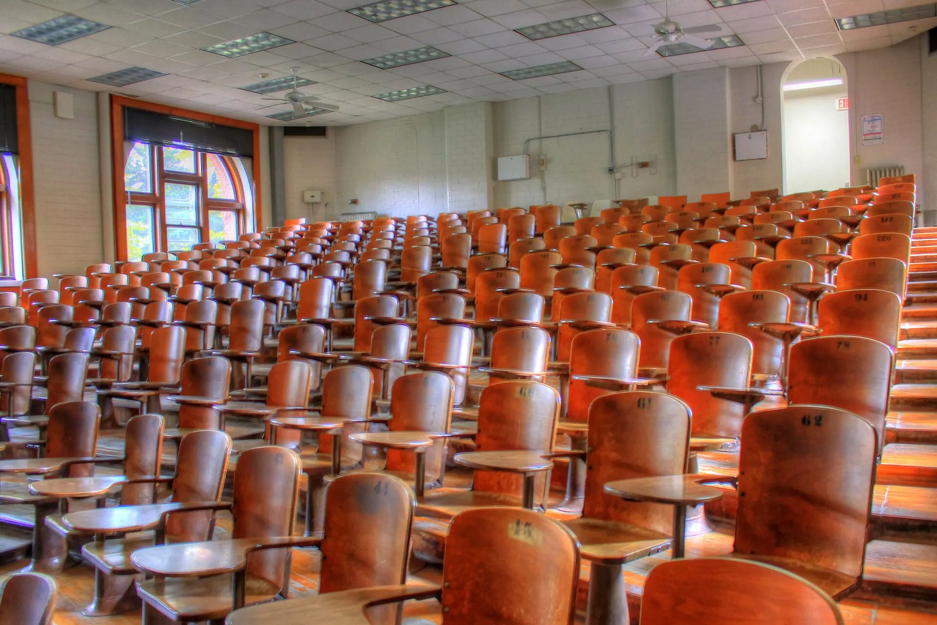 An empty tiered lecture hall with rows of numbered wooden tablet-arm seats rising toward the back, arched windows to one side and ceiling fans above