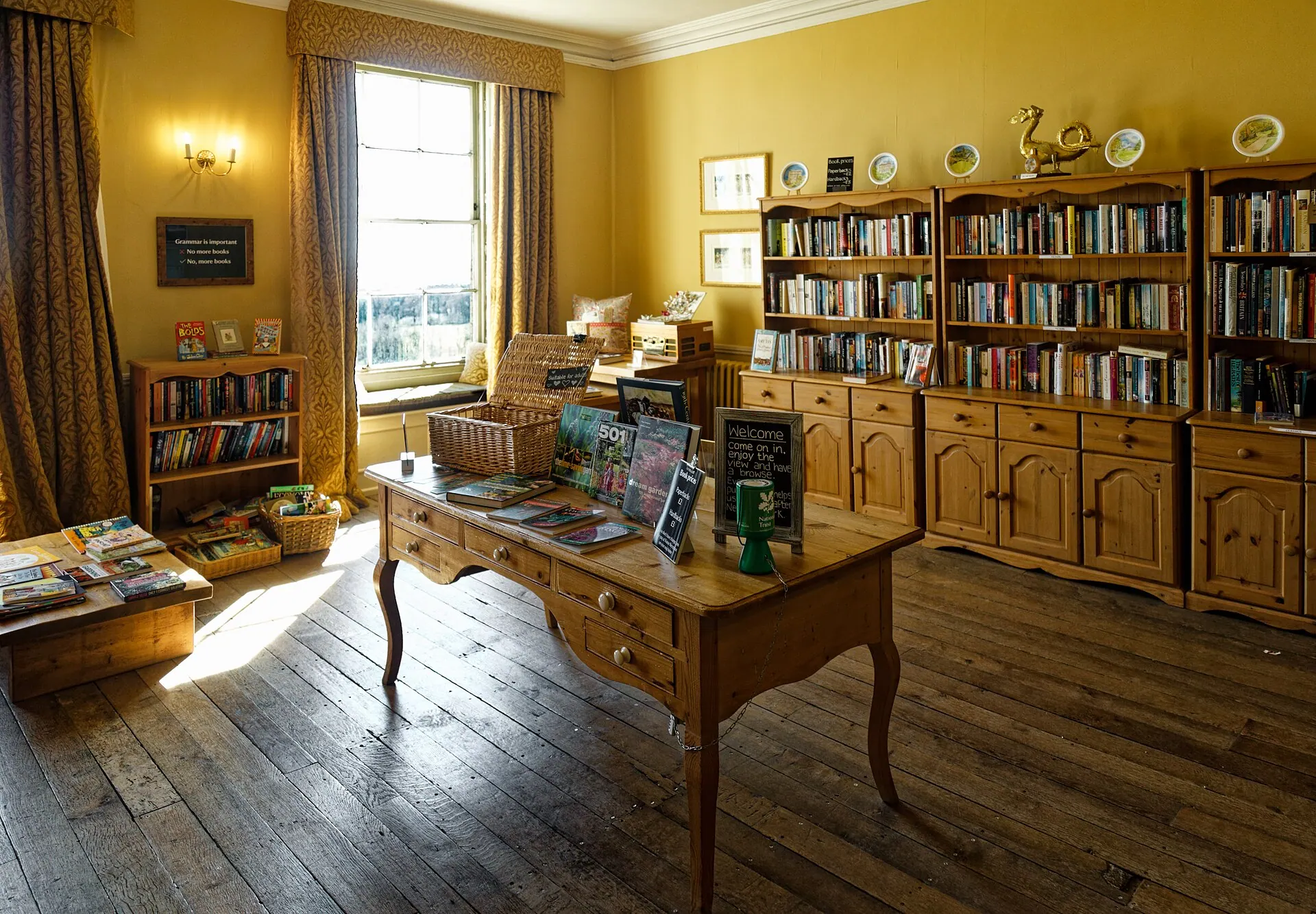 A sunlit book room with yellow walls, pine shelves packed with second-hand books, and a few volumes laid out on a wooden table in the middle