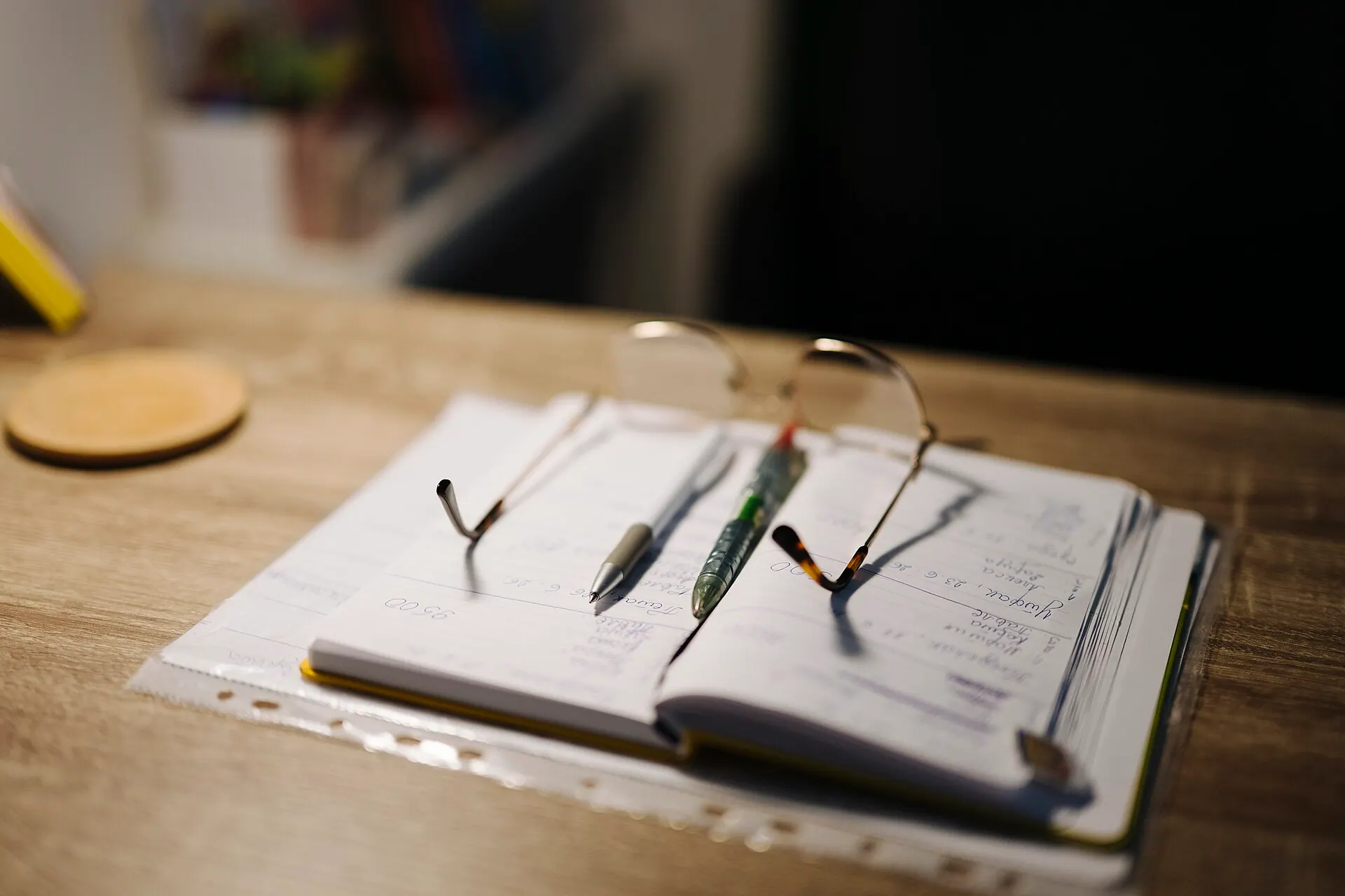 Cuaderno manuscrito abierto sobre un escritorio de madera, con dos bolígrafos y unas gafas apoyados en la página y luz lateral baja