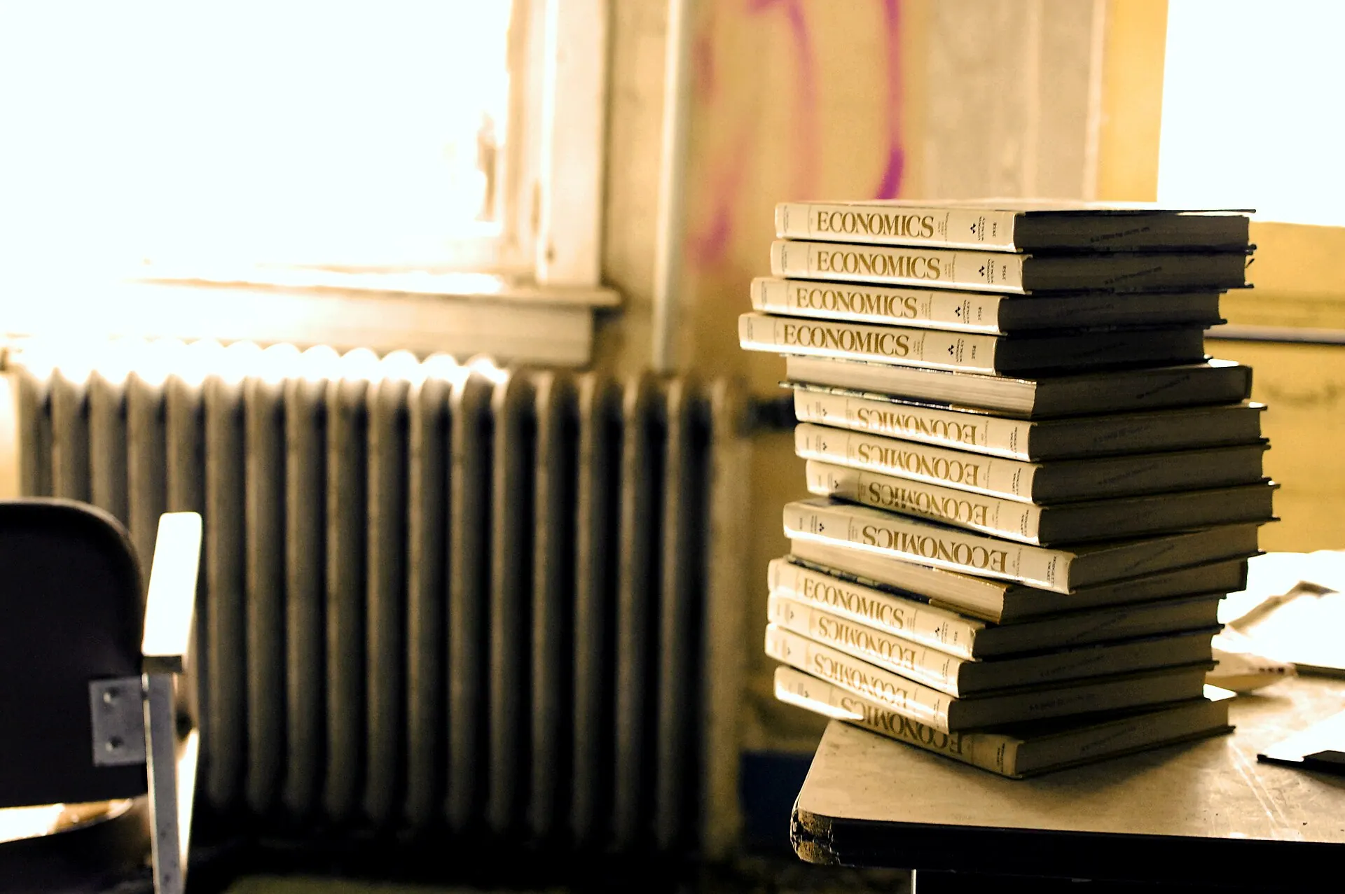 A tall stack of identical textbooks on a desk in a worn classroom, with a window and a cast-iron radiator behind