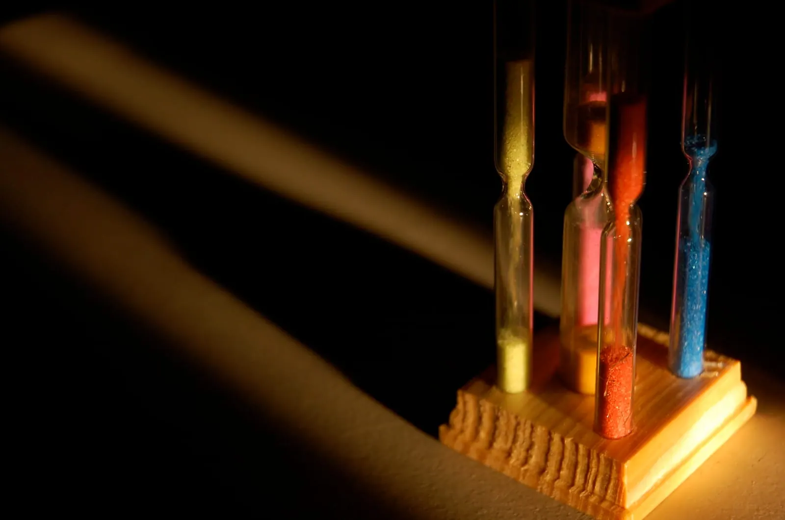 Quatro ampulhetas lado a lado numa base de madeira, cada uma com uma quantidade diferente de areia amarela, laranja, vermelha e azul, com luz lateral sobre fundo escuro
