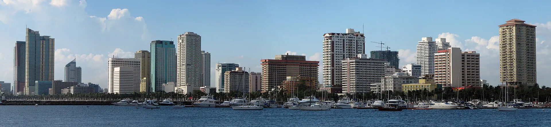The Manila waterfront skyline seen across the bay, a line of high-rises behind palm trees and yachts moored in the foreground