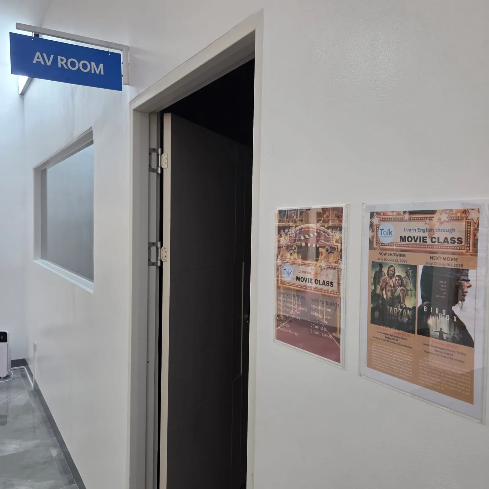 열린 문 위에 파란 AV ROOM 표지판이 걸린 어학원 복도, 벽에는 무비 클래스 포스터 2장