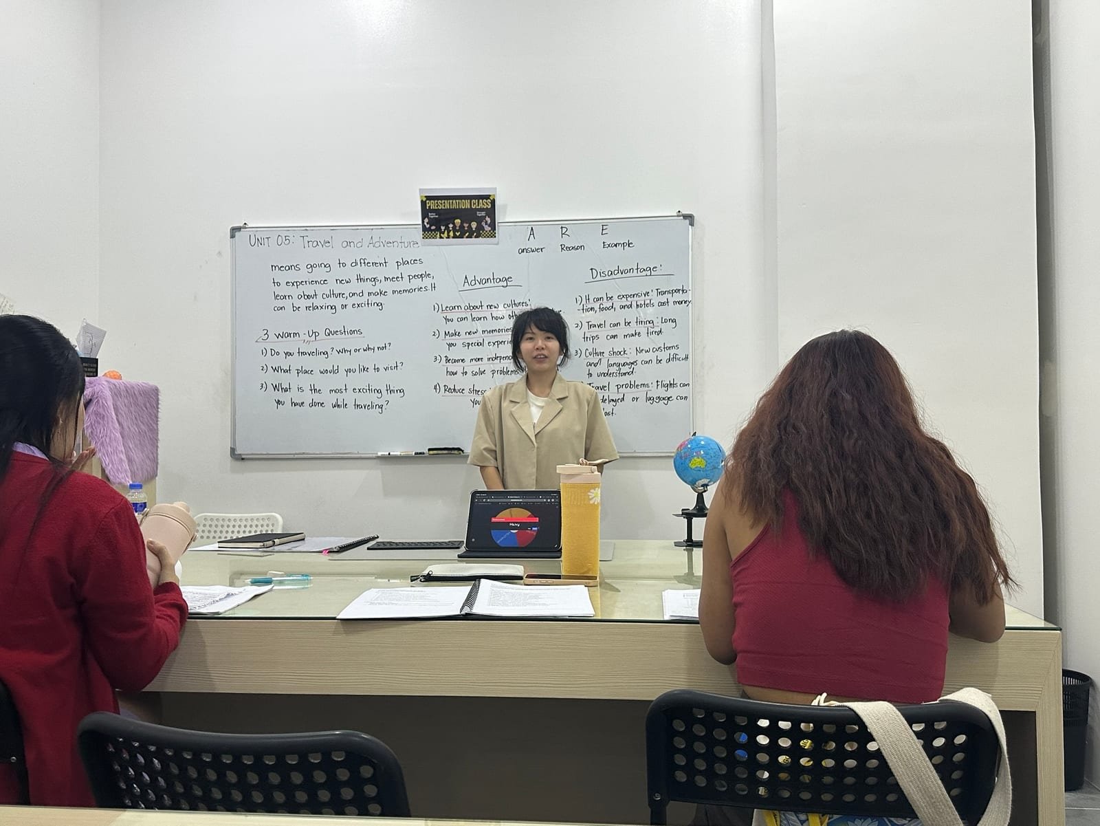 Aula em grupo pequeno, com a professora no quadro e os alunos de costas ouvindo