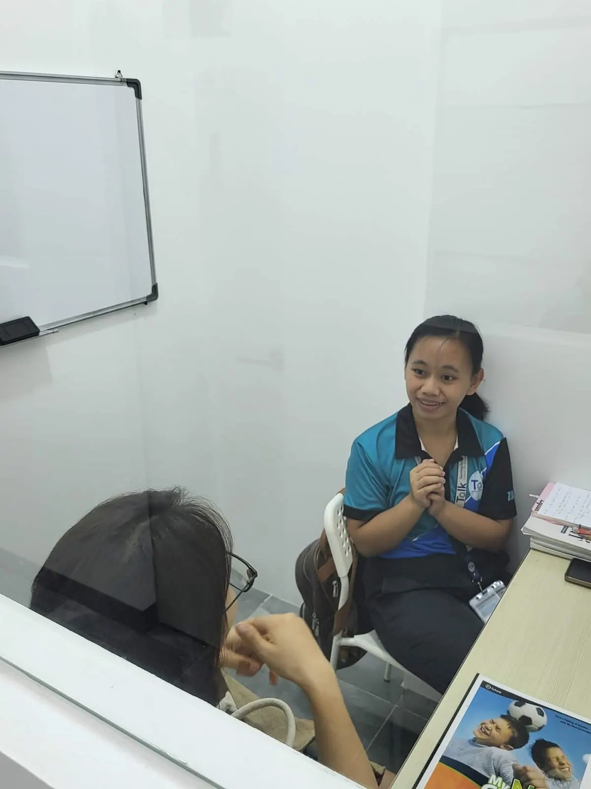 A one-on-one lesson in a booth-style classroom, teacher and student facing each other across a desk